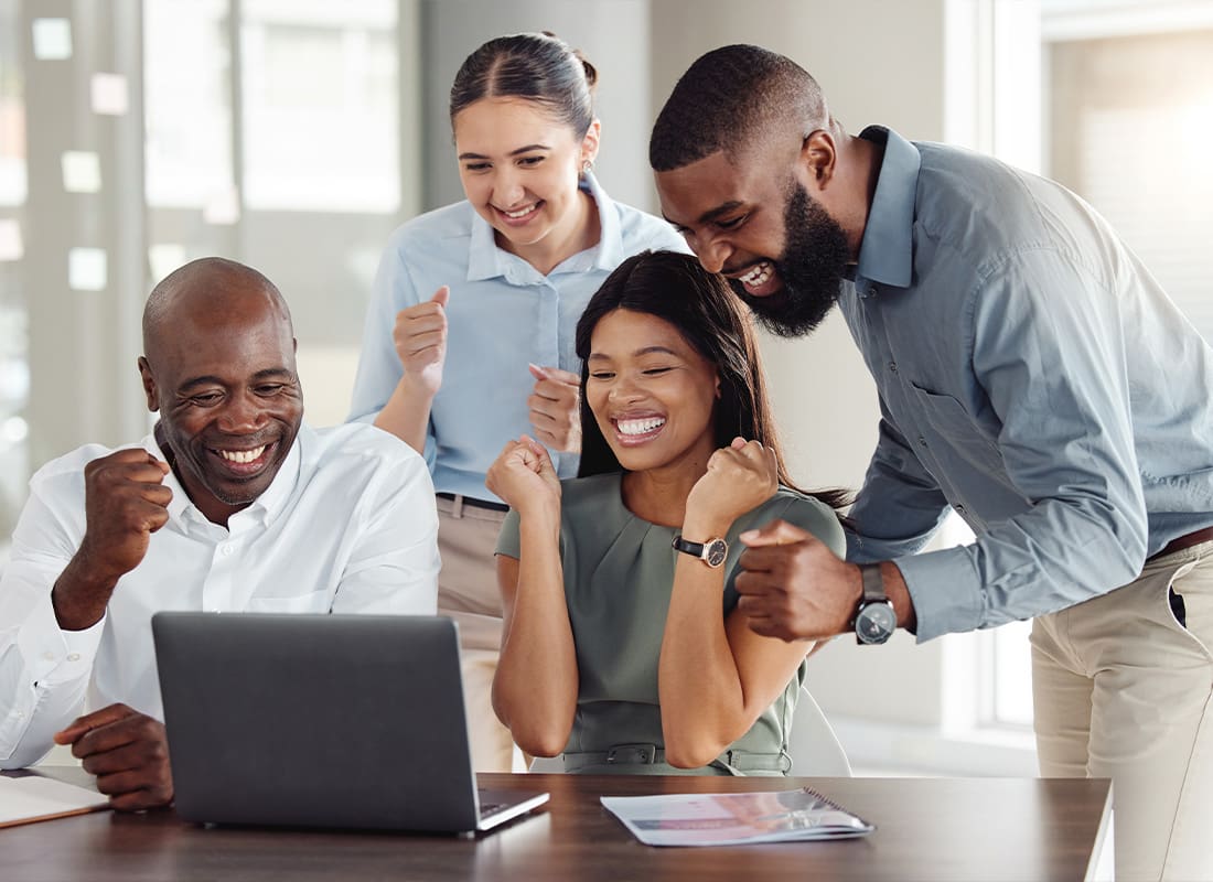 Employee Benefits - A Team of Employees Celebrating a Good Job While Sitting and Standing in Front of a Laptop at a Wooden Table